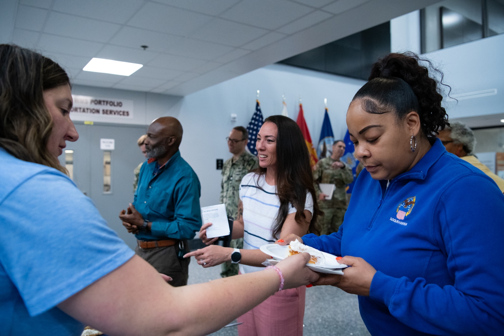 A woman wearing a blue shirt hands a piece of cake on a plate to another woman in a blue shirt as they stand inside a building lobby.