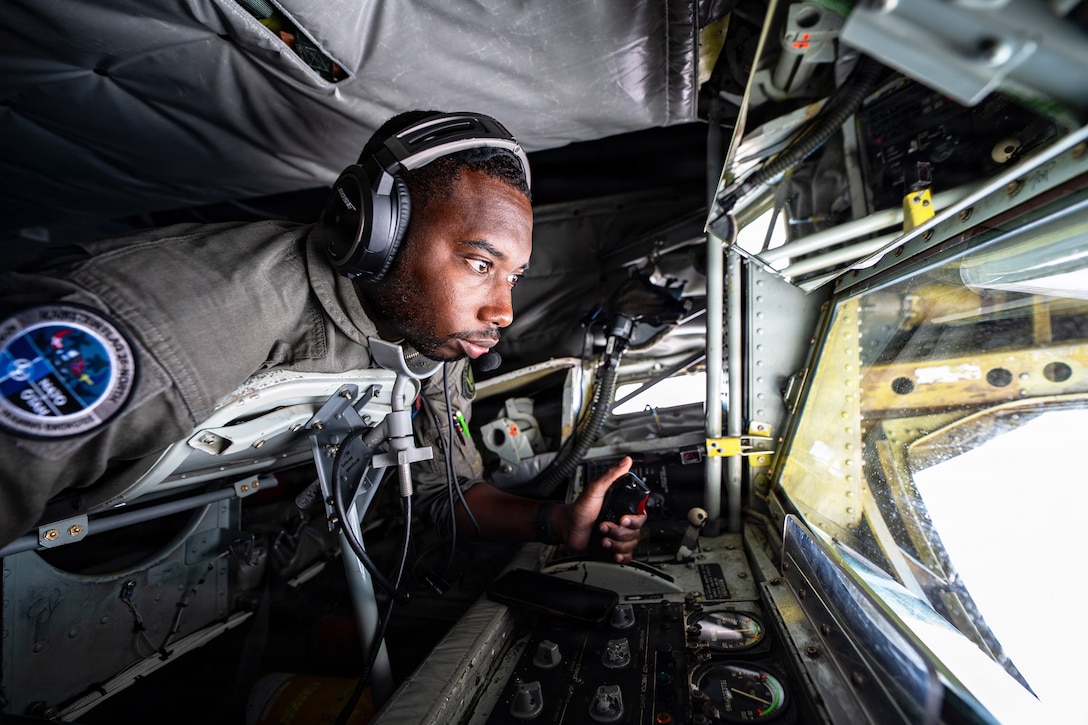An airman wearing a headset operates a remote-control stick in an aircraft's boom pod.