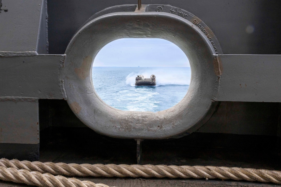 An air-cushioned landing craft transits a body of water during the day as seen through a ship’s porthole.