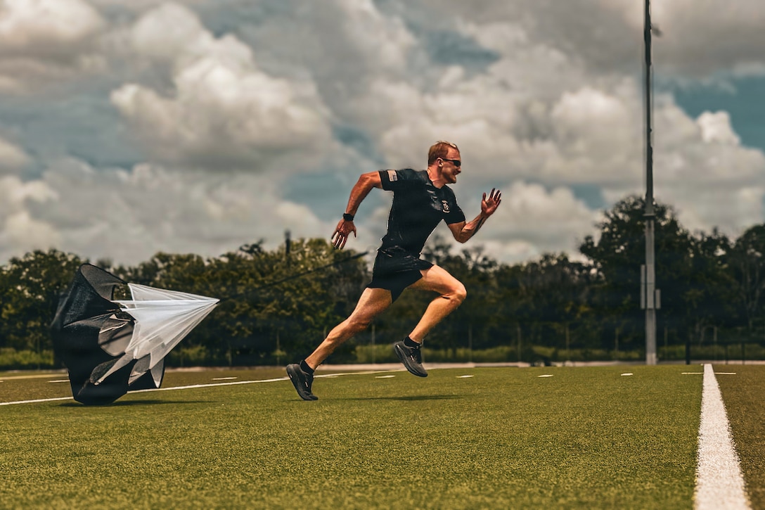A soldier runs with a small parachute attached to them on a turf during the day.