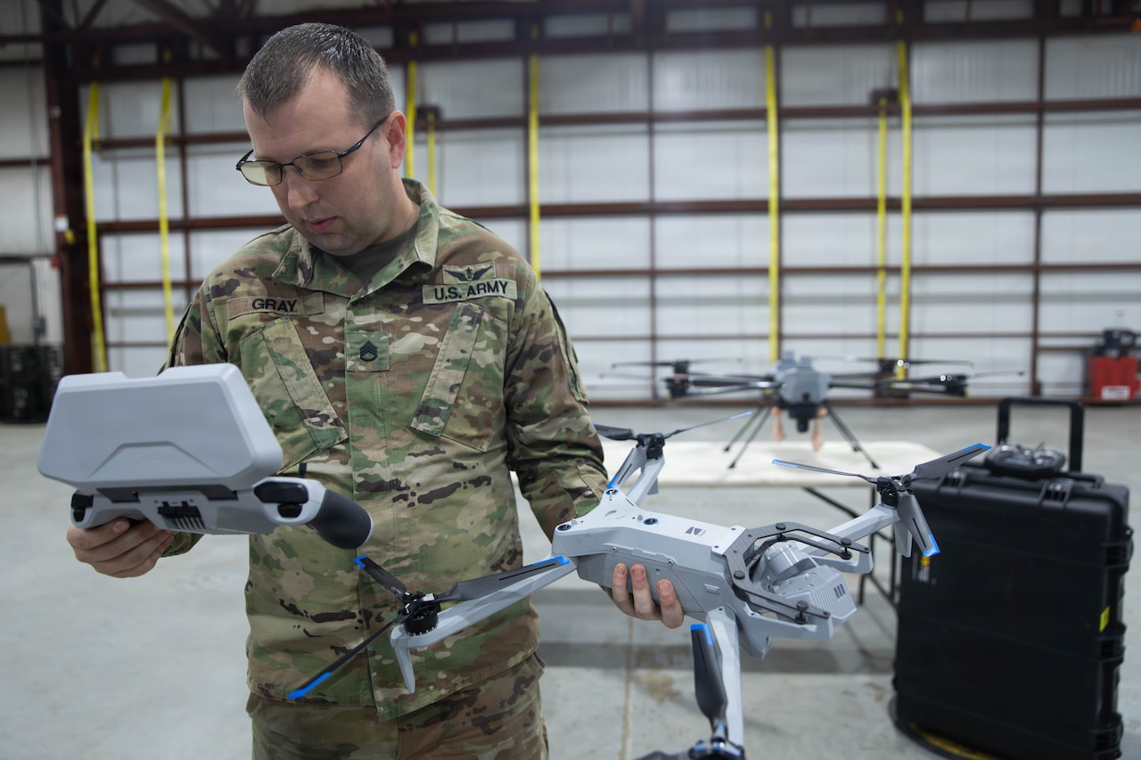 A man in a camouflage military uniform holds a drone in one hand and looks at a controller in the other.