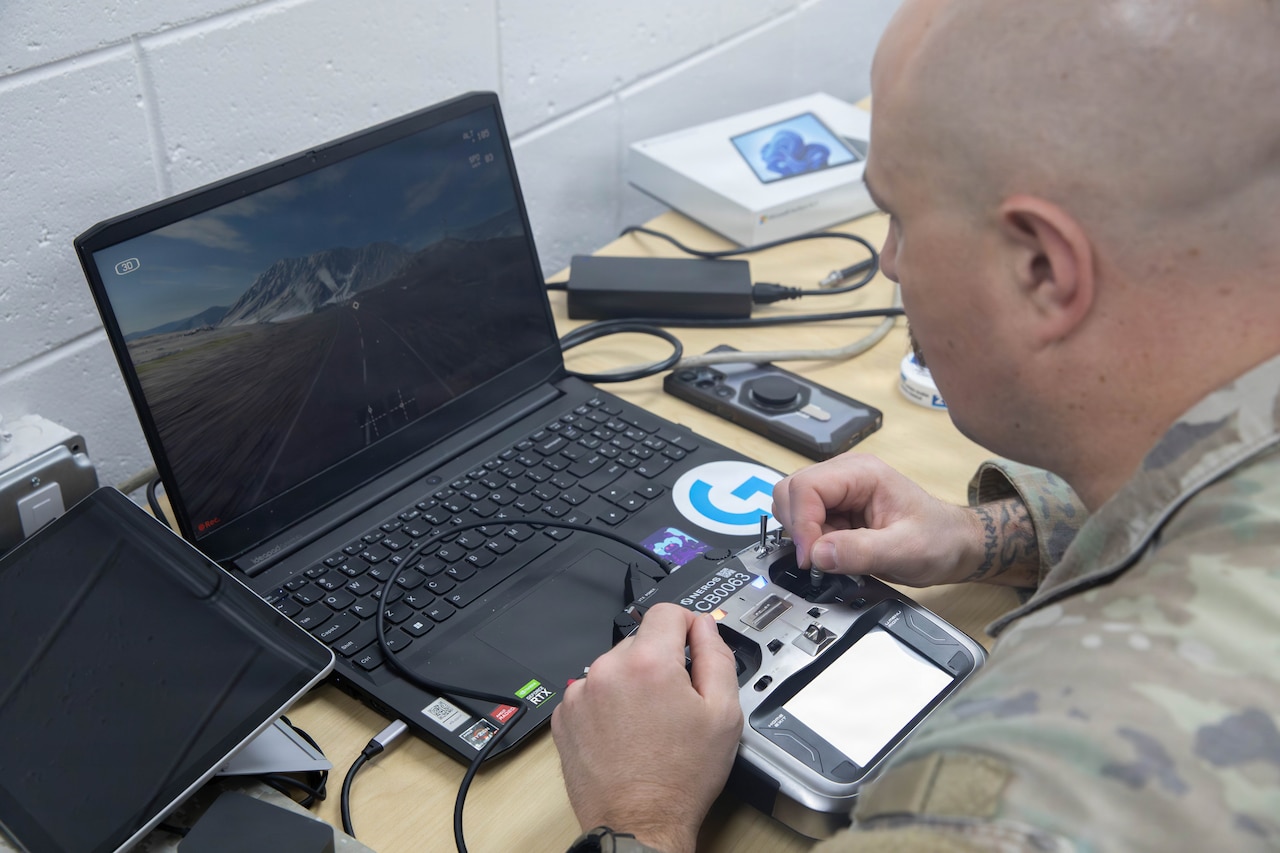 A man in a camouflage military uniform looks at a drone simulator on a laptop.