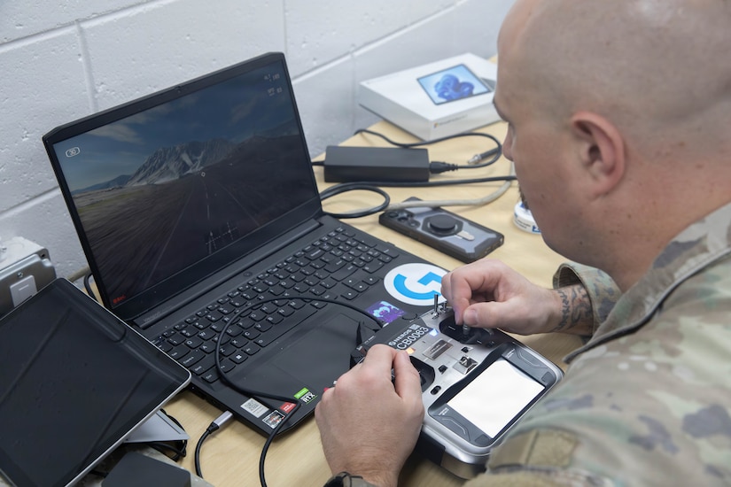 A man in a camouflage military uniform looks at a drone simulator on a laptop.