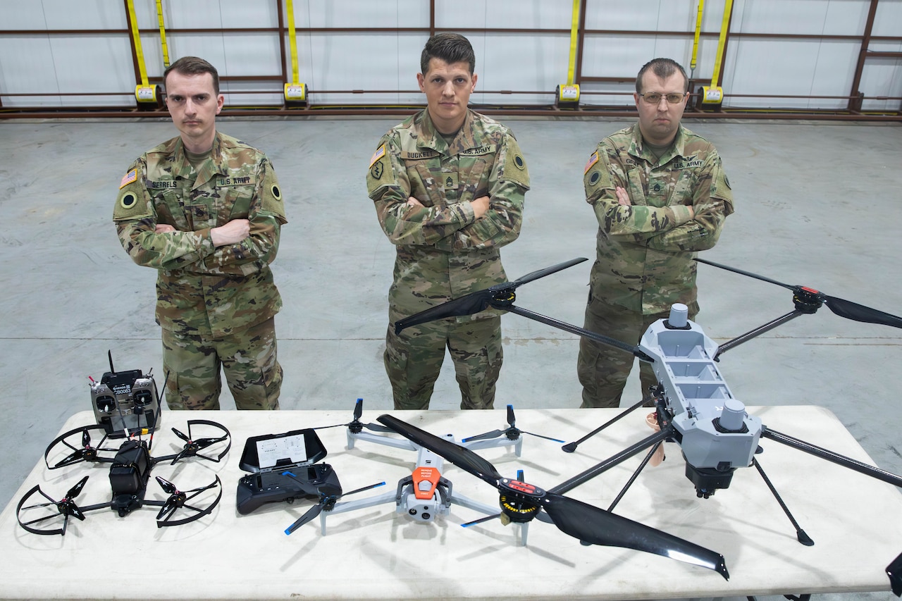 Three men in camouflage military uniforms stand and pose with arms folded behind a table filled with military drones.