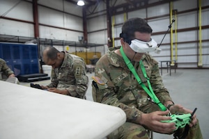 A man in a camouflage military uniform wears a headset and holds a remote control while seated in a warehouse. Another man in similar attire sits near him, holding a remote control.