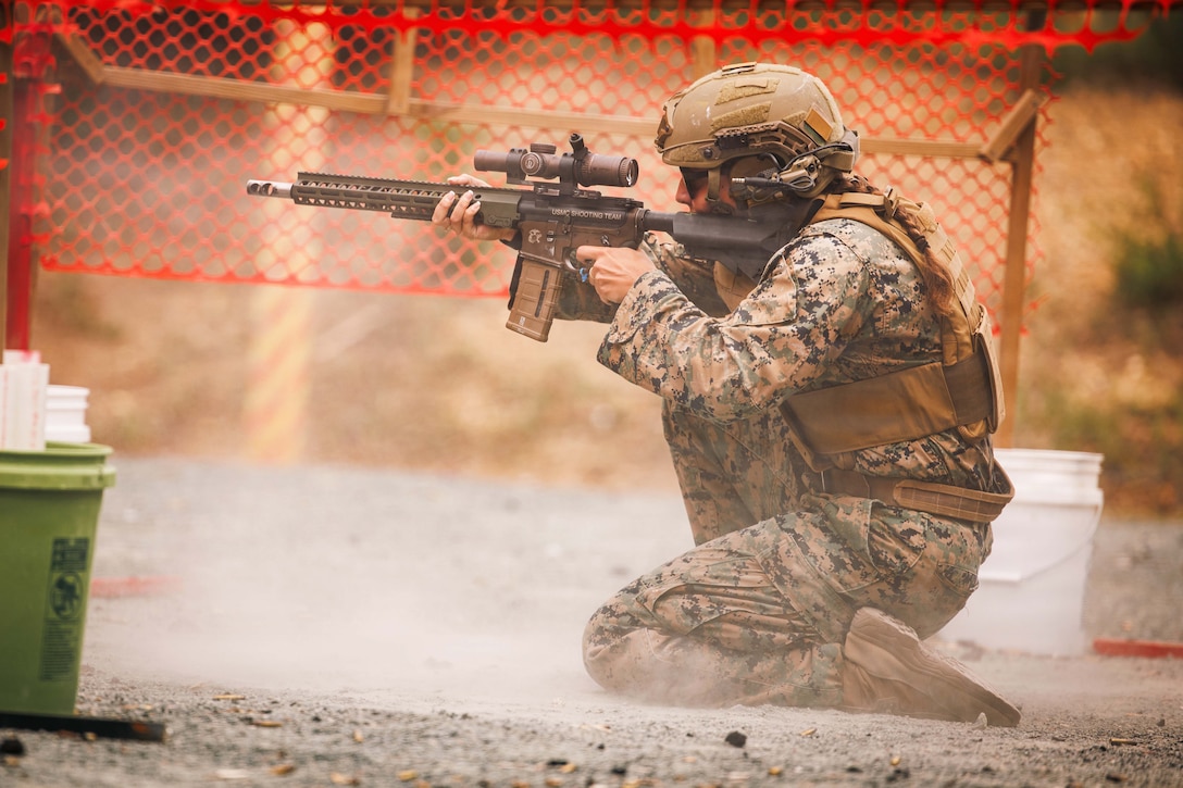 A Marine in tactical gear kneels in the dirt under an orange netted structure while aiming a rifle.