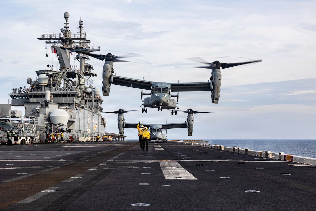 Sailors in yellow vests signal as an aircraft takes off from the flight deck of a ship at sea during the day.