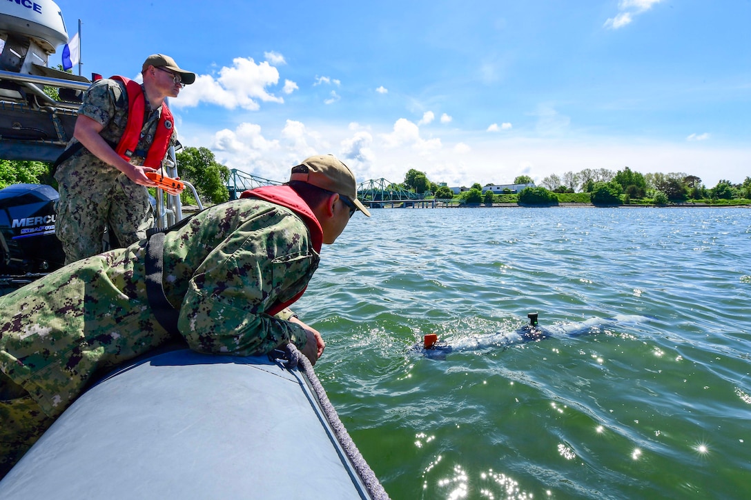 A sailor uses an orange remote control as another sailor watches an unmanned underwater vehicle transit murky waters from a boat during the day.