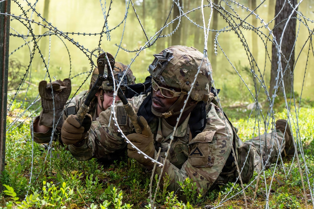 Soldiers in tactical gear lying in a wooded area use wire cutters to cut barbed wire during the day.