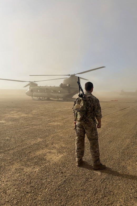 A man wearing a camouflage military uniform stands with a weapon strapped around his shoulder in a dirt field as a helicopter in front of him kicks up dust. There is another helicopter in the distance.