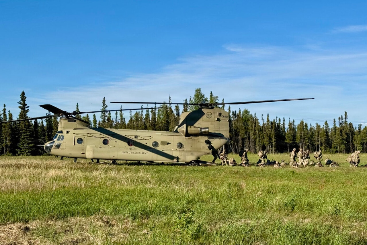 Soldiers wearing combat gear disembark a helicopter outdoors in a field lined by trees.