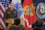 An Army Lieutenant General stands at a podium with military flags in the background.