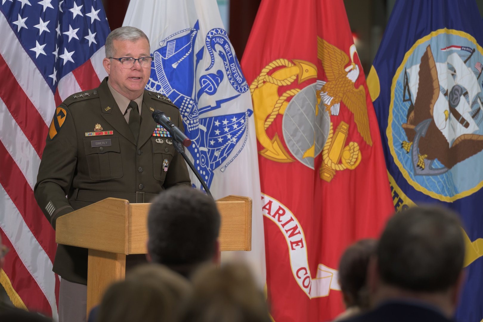 An Army Lieutenant General stands at a podium with military flags in the background.