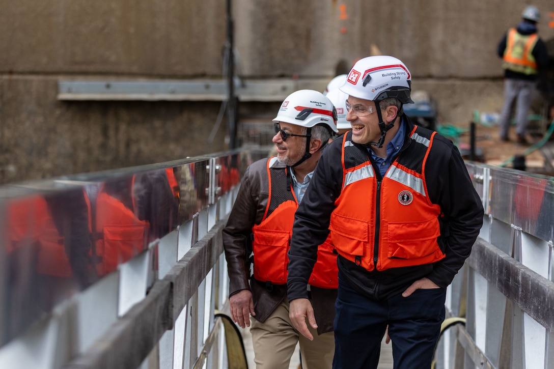 Two smiling men in hard hats and orange life vests walk and talk on a narrow metal walkway.