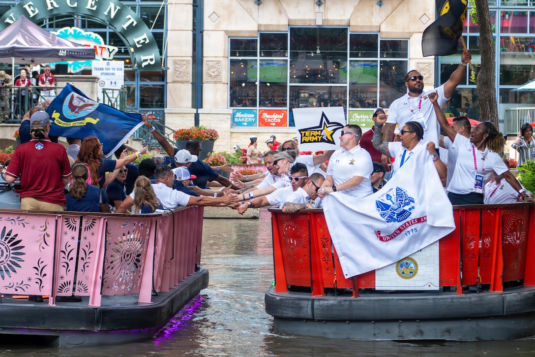 Competitors from different services shake hands while passing each other in boats in a river in front of a building.