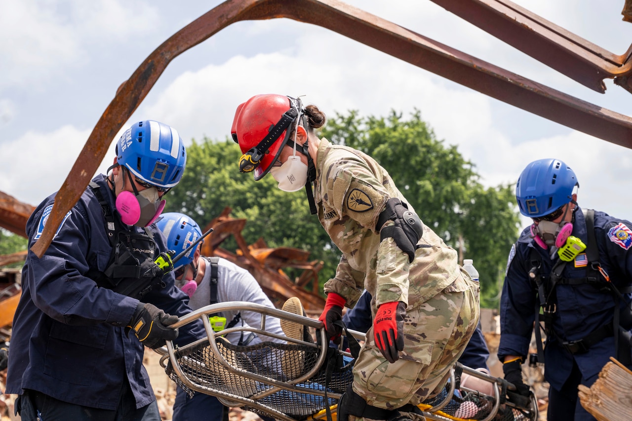 A soldier and fellow responders wearing protective gear carry a metal gurney near a tree under a blue sky.