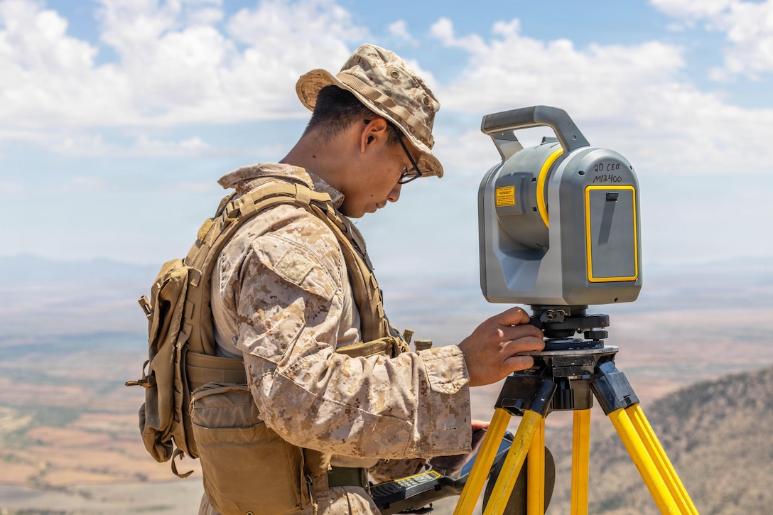 A Marine sets up survey equipment on a yellow tripod on mountainous terrain under a blue sky.