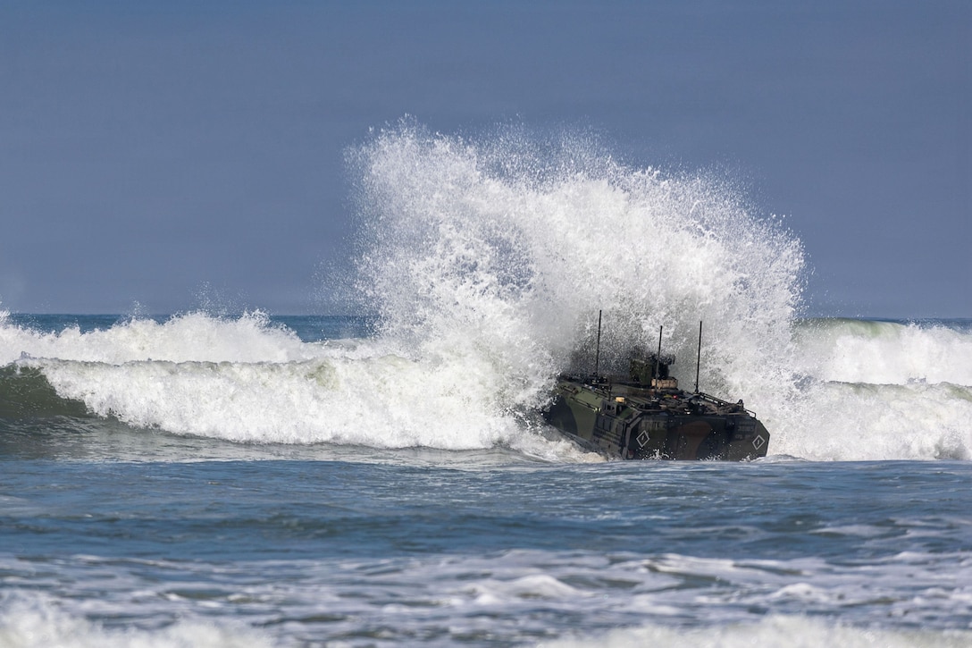An amphibious combat vehicle makes a large splash in a body of water under a blue sky.