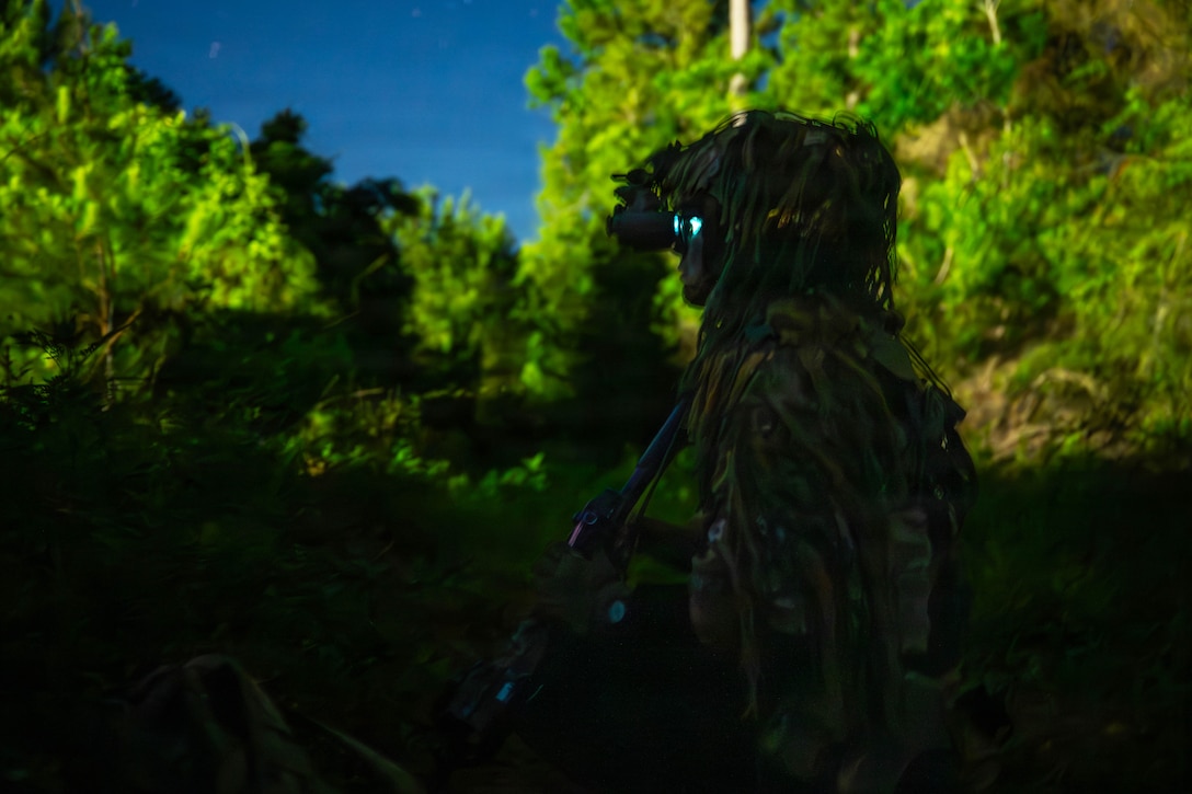 A soldier in a camouflage uniform and a helmet covered in foliage stands in the shade and looks through binoculars while holding a weapon in the brush.