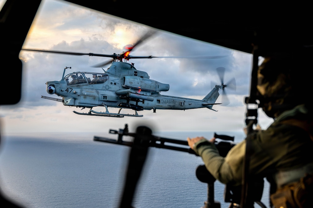 A helicopter is in the sight line of a Marine in another helicopter in the air over the sea.