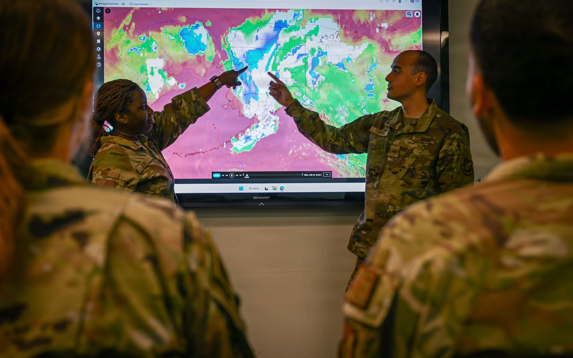 Two Airmen point to satellite imagery of Alaska shown on a large screen inside a conference room