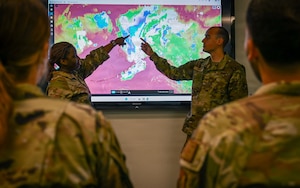 Two Airmen point to satellite imagery of Alaska shown on a large screen inside a conference room