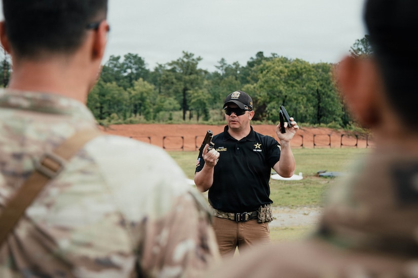 A man wearing casual attire and a baseball cap holds up a pistol and two ammunition magazines as he talks to people dressed in camouflage military uniforms in the foreground.