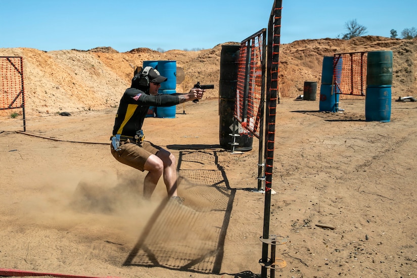 A man wearing casual attire fires a pistol on a training range as dust is kicked up around his feet; there are mounds of dirt and obstacles in the background.