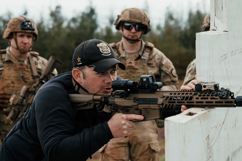 A man wearing casual attire and a baseball cap aims a rifle while propping it on top of a wooden post; there are three men dressed in camouflage military uniforms observing in the background.