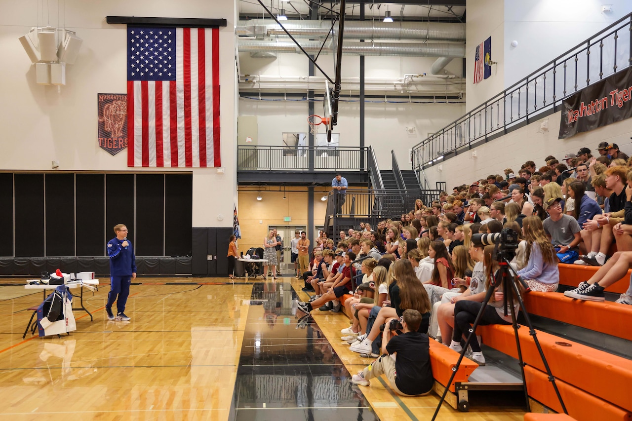 A man wearing athletic attire speaks to dozens of kids seated on the bleachers inside a gym; there is a large American flag hanging from a wall in the background.