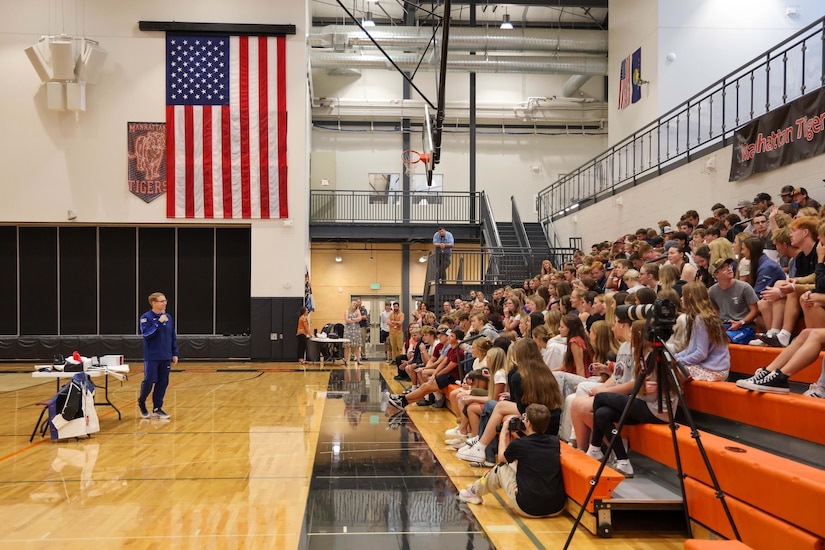 A man wearing athletic attire speaks to dozens of kids seated on the bleachers inside a gym; there is a large American flag hanging from a wall in the background.