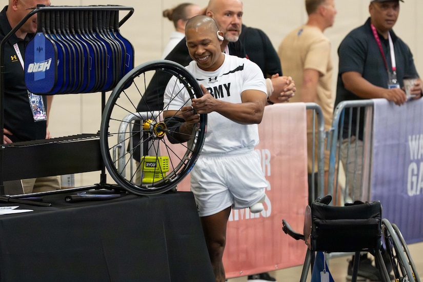 A man in athletic attire stands on one leg next to a table and holds a wheelchair wheel.