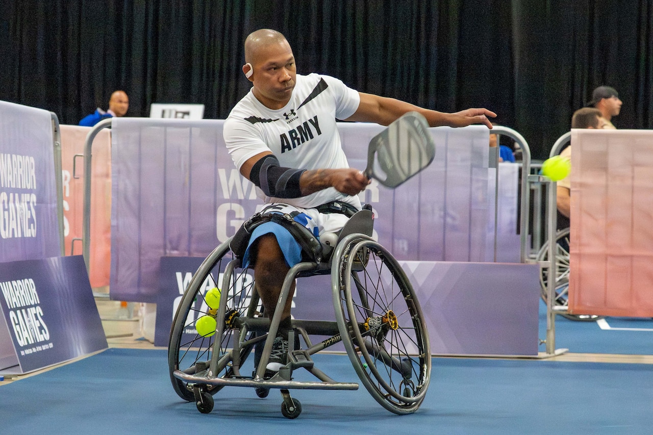 A man in athletic attire sits in a wheelchair and swings a pickleball racket.