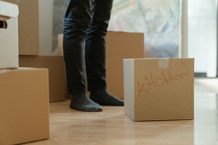 A person's legs in dark pants and socks in a kitchen, surrounded by cardboard boxes. "Kitchen" is written on one of the boxes.