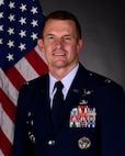 Col Scott Mills poses for an official photo with a U.S. flag behind him with a grey background.