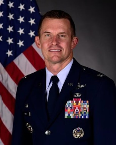 Col Scott Mills poses for an official photo with a U.S. flag behind him with a grey background.