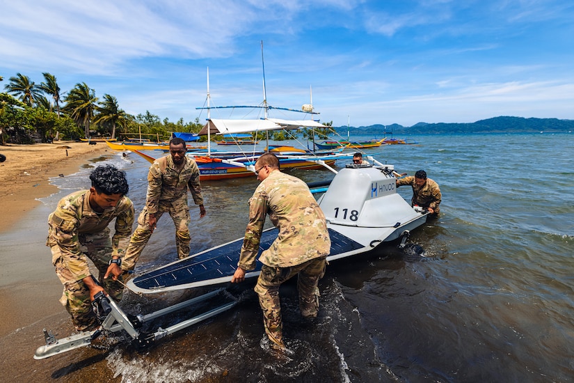 Five men wearing camouflage military uniforms push an unmanned surface vessel out of the water and onto a trailer while standing on a beach; there are several small boats in the background.