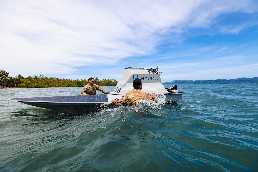 Two men wearing camouflage military uniforms stand in waist-deep water as they inspect an unmanned surface vessel; there are trees and mountains in the background.