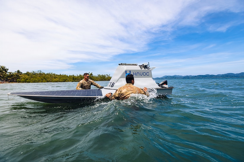 Two men wearing camouflage military uniforms stand in waist-deep water as they inspect an unmanned surface vessel; there are trees and mountains in the background.