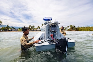 Two men wearing camouflage military uniforms stand in waist-deep water as they inspect an unmanned surface vessel; there are houses and trees in the background.