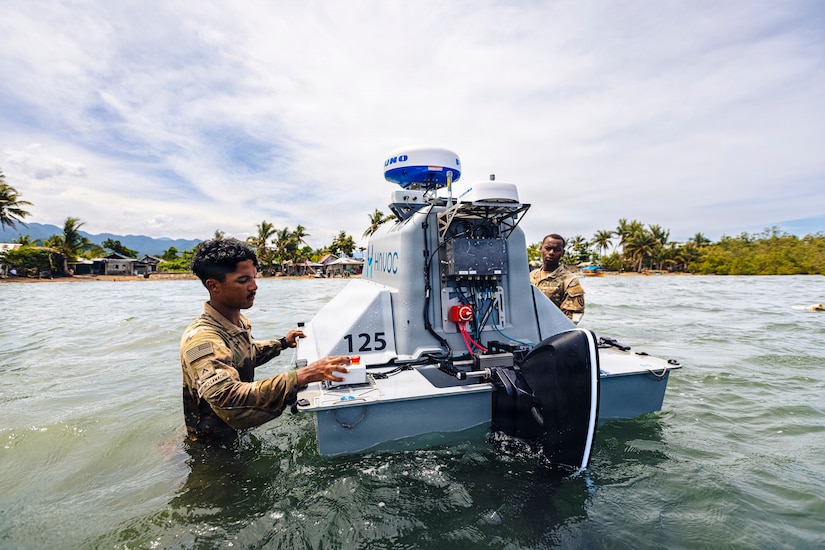 Two men wearing camouflage military uniforms stand in waist-deep water as they inspect an unmanned surface vessel; there are houses and trees in the background.