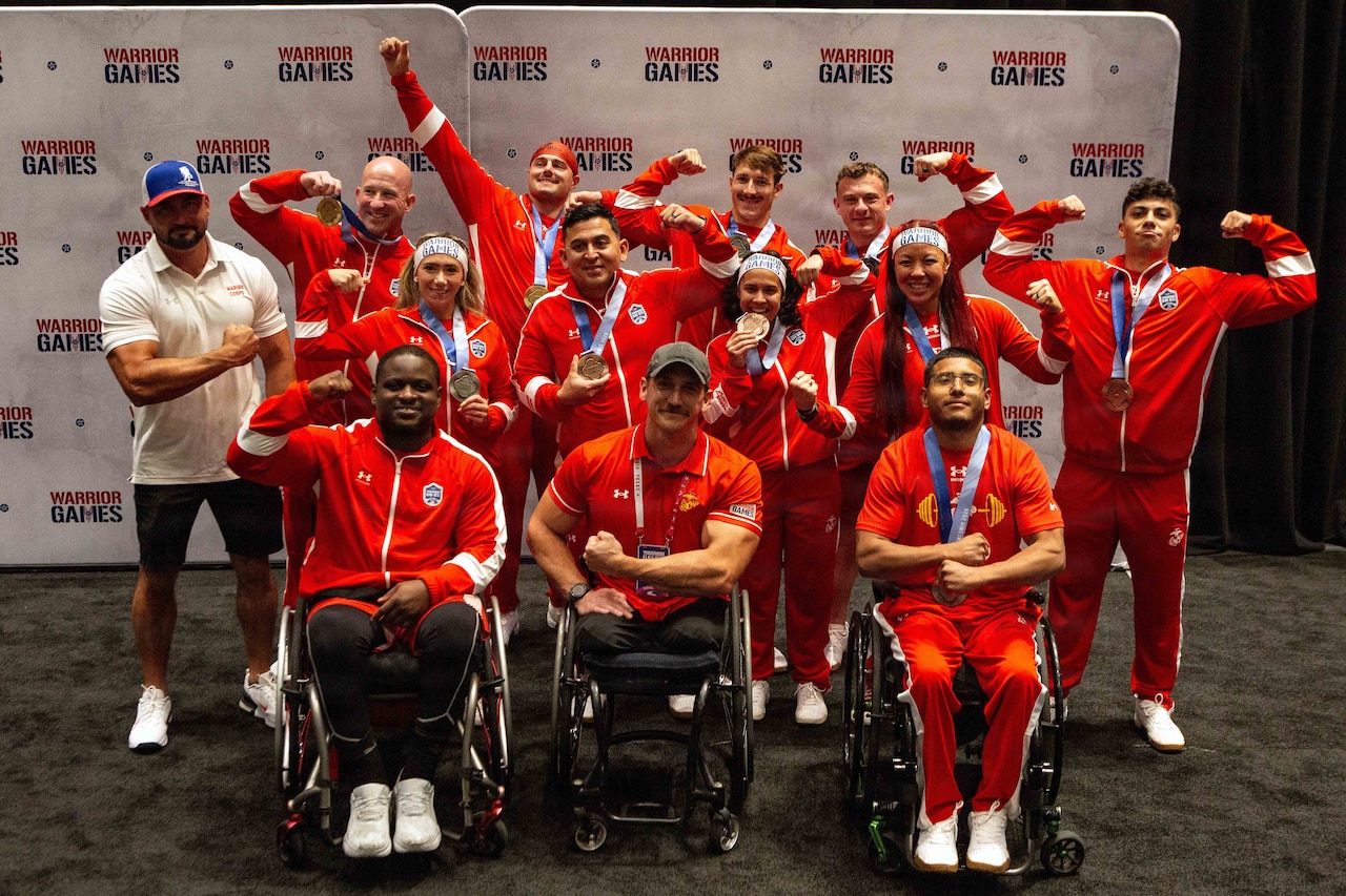 A dozen people in athletic attire pose for a photo with medals around their necks; most of them are either holding up the medal or flexing their muscles. The three men in the front row are sitting in wheelchairs.