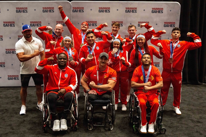A dozen people in athletic attire pose for a photo with medals around their necks; most of them are either holding up the medal or flexing their muscles. The three men in the front row are sitting in wheelchairs.