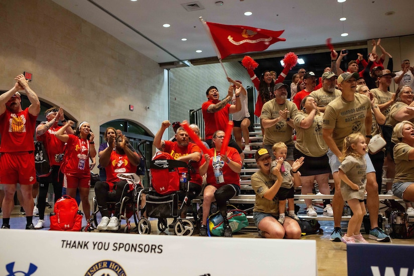 Dozens of men, women and children cheer from the bleachers of a gym; a man in the background is waving the Marine Corps flag.
