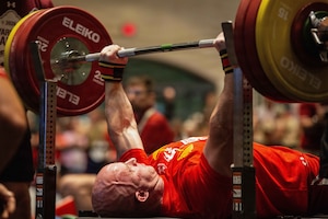 A close-up of a man in athletic attire as he prepares to bench press a large amount of weight during a competition, while other people in similar attire observe.