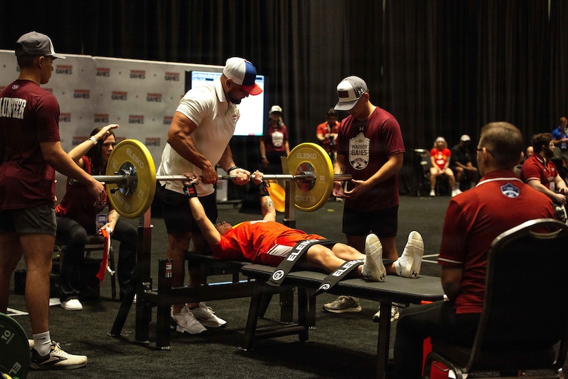 A woman in athletic attire prepares to bench press during a competition while her legs are strapped to the bench; there are several people in similar attire spotting her or observing.
