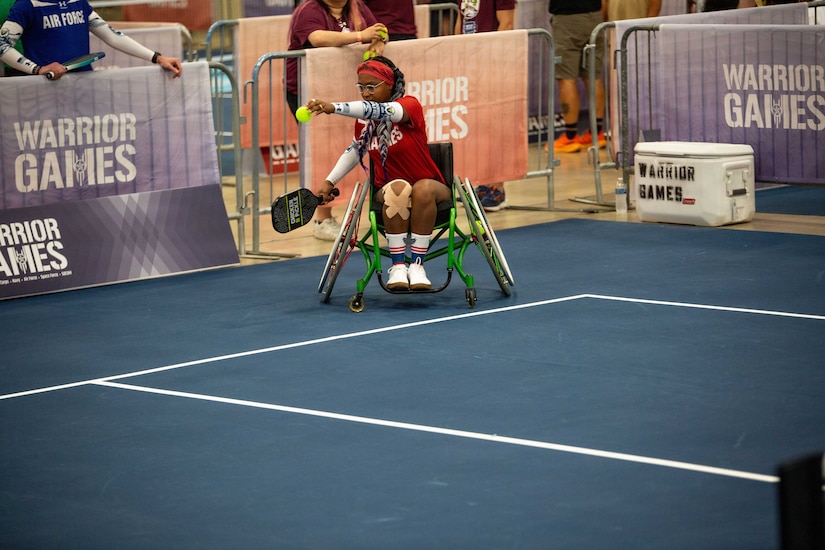 A woman in athletic attire sits in a wheelchair as she prepares to serve the ball during an indoor pickleball game.