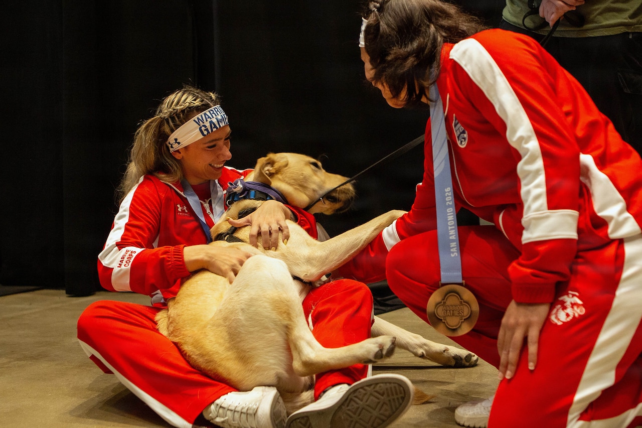 A woman in athletic attire pets a dog as the dog sits in her lap; another woman in similar attire with a medal around her neck kneels while also petting the dog.