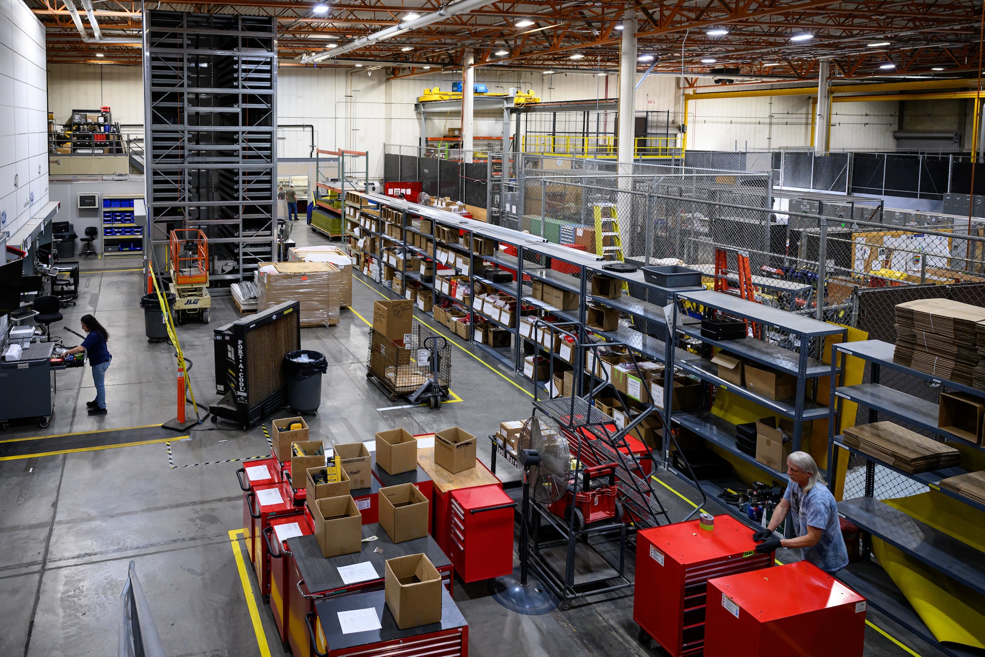 A high-angle view of the 309th Maintenance Support Group Tools Flight warehouse at Hill Air Force Base, showing rows of shelving, boxes, and specialized equipment as workers organize inventory on the floor.