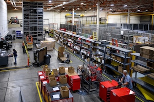 A high-angle view of the 309th Maintenance Support Group Tools Flight warehouse at Hill Air Force Base, showing rows of shelving, boxes, and specialized equipment as workers organize inventory on the floor.